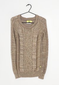 Stickad tröja i ljusbrunt med texturerade kabelmönster och små dekorationer. Har en ribbad krage och muddar, hängande på en svart krok.