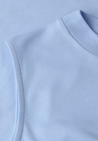 Gros plan sur un tissu bleu clair sans manches avec un col montant, montrant une texture lisse et des coutures détaillées sur l'épaule et l'encolure.