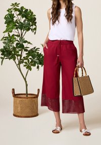 Červené zkrácené kalhoty s výraznou krajkou, bílé bezramínkové topy a tkaná hnědá kabelka. Bílá sandály dokončují outfit.