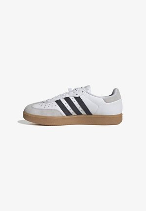 Zapatilla blanca con rayas negras, puntera y talón de ante gris, y suela de goma color natural, mostrada de perfil sobre fondo blanco.
