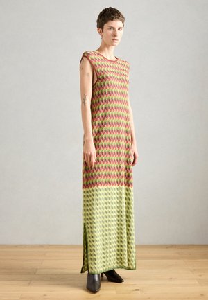 Person med kort hår iført en ærmeløs, mønstret maxi-kjole i røde, grønne og gule nuancer, stående på et trægulv mod en grå væg.