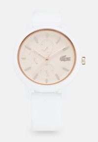Reloj de silicona blanca con un bisel redondo dorado rosa, esfera crema, tres subesferas y un logotipo de cocodrilo; presenta marcadores de hora y agujas luminiscentes.