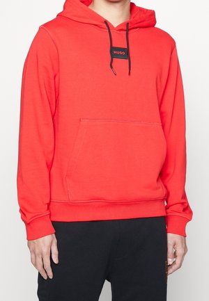 Person iført en klar rød hættetrøje med forlommer og sort "HUGO" logo patch, kombineret med sorte bukser mod en hvid baggrund.