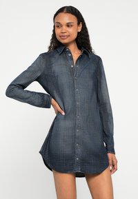 Φόρεμα πουκάμισο από τζιν σε σκούρο μπλε με κουμπιά μπροστά, μακριά μανίκια και στρογγυλεμένο τελείωμα. Διαθέτει διακριτικό καρό μοτίβο και άνετη εφαρμογή.