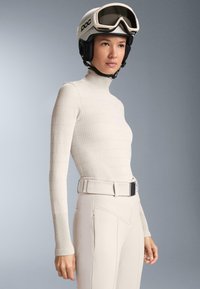 Jasnobeżowa prążkowana bluzka z długim rękawem i wysokim kołnierzem w połączeniu z jasnobeżowymi spodniami oraz białym kaskiem narciarskim z goglami.