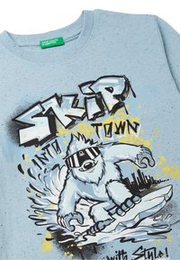 Světle modré bavlněné tričko s grafikou kresleného yetiho se slunečními brýlemi a skateboardem, doplněné černým a žlutým rozstřikovaným vzorem.