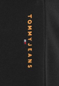 Μαύρο ύφασμα με το κείμενο «TOMMY JEANS» κεντημένο κάθετα σε πορτοκαλί χρώμα, με ένα μικρό τρίχρωμο λογότυπο σε μπλε, άσπρο και κόκκινο.