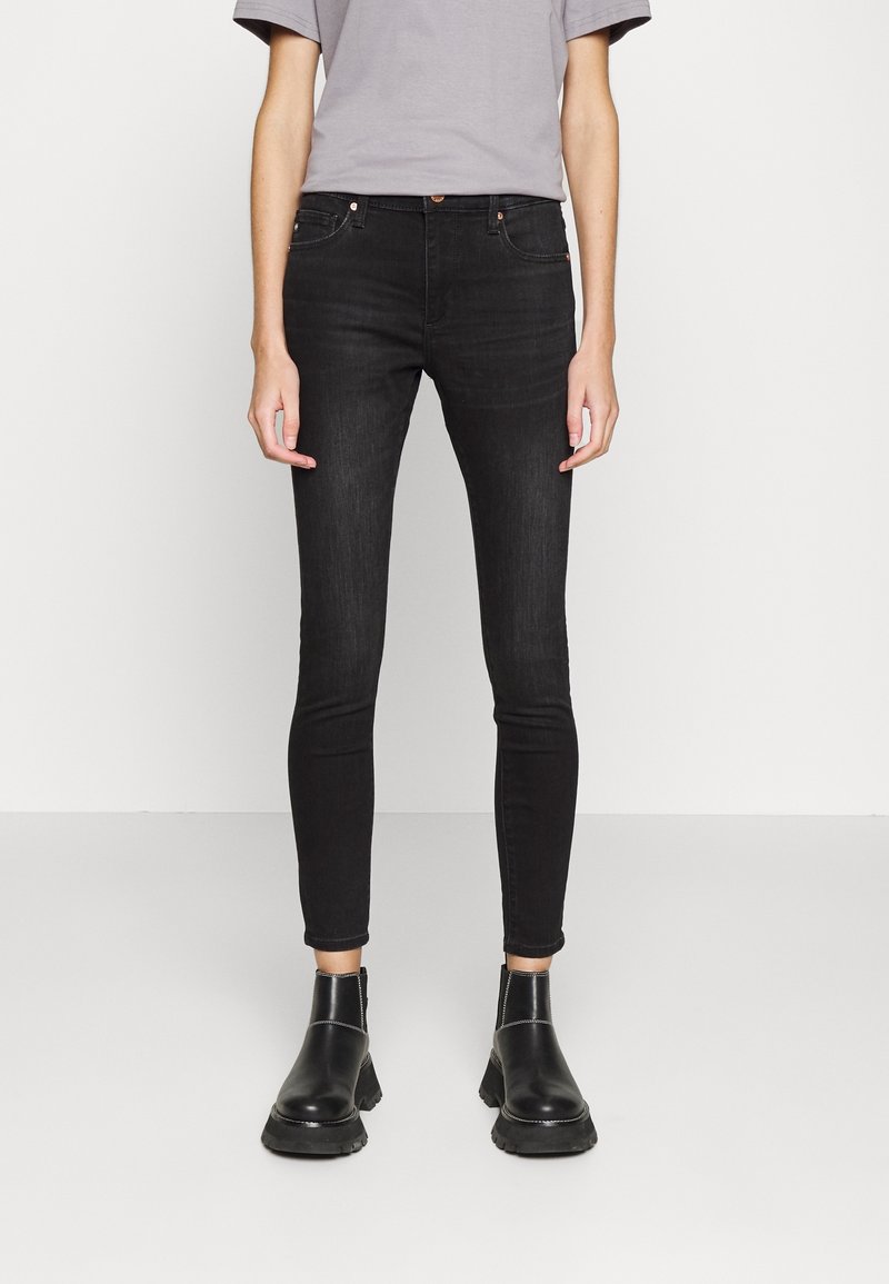 Jeans negros ajustados de mezclilla, con cinco bolsillos, cierre de botón y diseño minimalista. Llevados con botas negras hasta el tobillo.