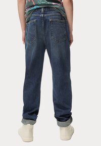 Jeans de mezclilla azul oscuro con puños enrollados, que cuentan con un diseño de cinco bolsillos y una cremallera. La parte trasera tiene dos bolsillos cosidos y una costura tipo yugo.