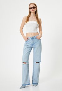 Top corto blanco con fruncidos y bordes con volantes, combinado con jeans de pierna ancha en azul claro con efecto desgastado, que presentan rotos en las rodillas y un dobladillo deshilachado.