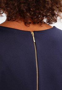 Marinblå klänning med texturerat tyg, båt-ringning och en synlig guldfärgad dragkedja bak för stängning.