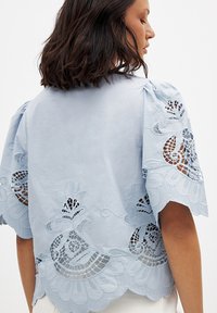 Lichtblauwe blouse met opgeblazen mouwen, bloemenkantaccenten langs de zoom en mouwen, en een zachte, gestructureerde stof.