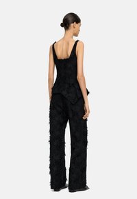 Czarny zestaw o teksturowanej powierzchni, składający się z dopasowanej bluzki z dekoltem z tyłu oraz spodni o szerokich nogawkach, oba elementy mają ozdobne frędzle. Gładkie czarne obuwie.