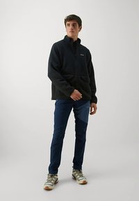 Črna flis jakna z visokim ovratnikom in zadrgo, kombinirana s temno modrimi hlačami in bež pohodnimi čevlji z sivimi in zelenimi poudarki.