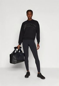 Melns DKNY sporta džemperis ar reljefu logotipu, saskaņots ar melnām legingām. Modele tur melnu sporta somu, valkājot melnas sporta kurpes.