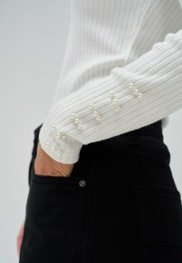 Witte geribde trui met lange mouwen, met parelversieringen op de manchetten. Draagt samen met donkere jeans. Focus op textuur en details.