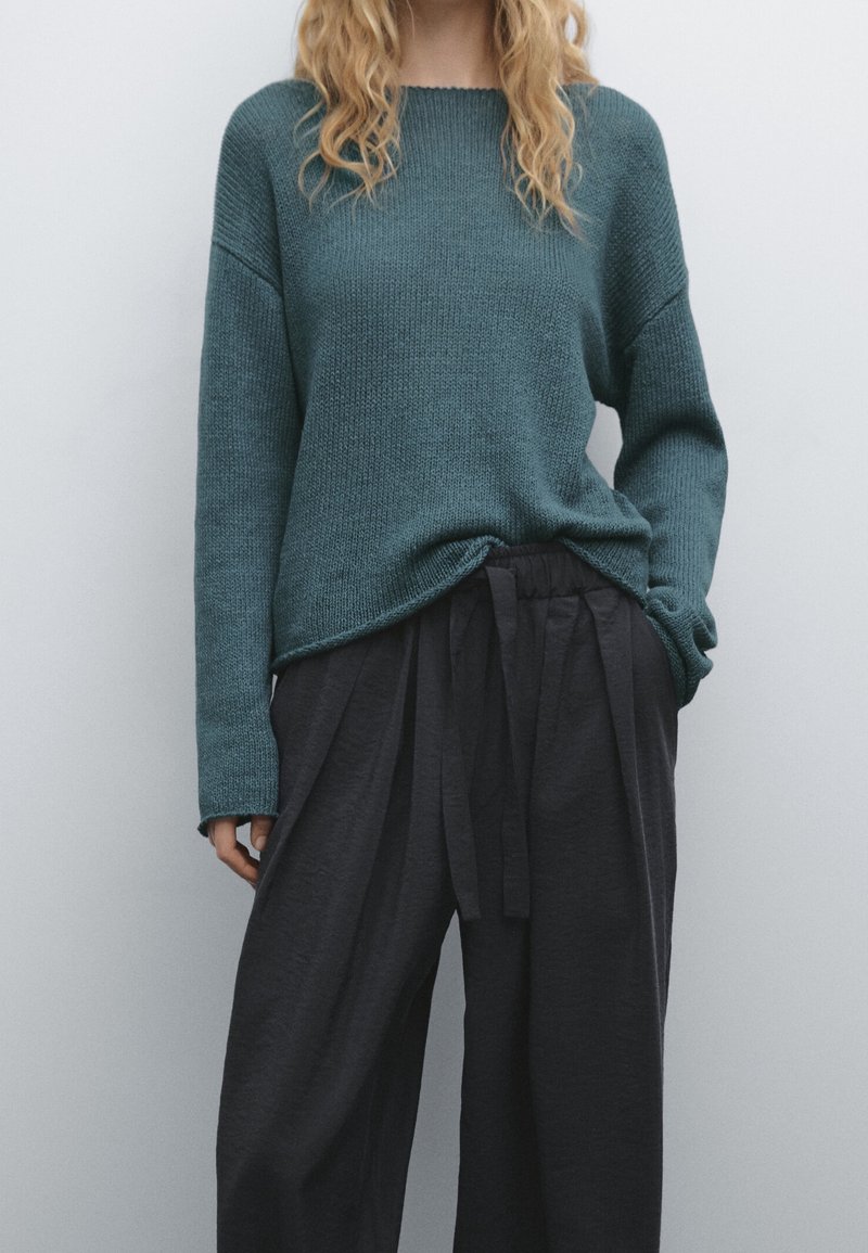 Personne portant un pull en maille bleu sarcelle à manches longues, rentré dans un pantalon ample noir à cordon, debout devant un fond uni.