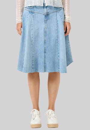 Średniej długości jasnoniebieska spódnica dżinsowa z surowym wykończeniem, noszona z białą koronkową bluzką z długim rękawem i białymi sneakersami na platformie na szarym tle.