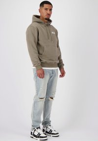 Olive green hoodie with front pocket, drawstring hood, and white logo. Worn with light blue ripped jeans and black-and-white sneakers.