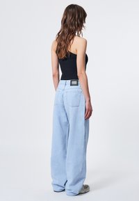 Hellblaue weit geschnittene Jeans mit entspannter Passform, versehen mit einem schwarzen Logo-Patch auf der Rückseite und vier Taschen. Glatte Denim-Oberfläche.