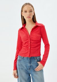 Rød, struktureret langærmet skjorte med krave, knaplukning foran og draperingsdetaljer. Sammen med blå denimjeans.