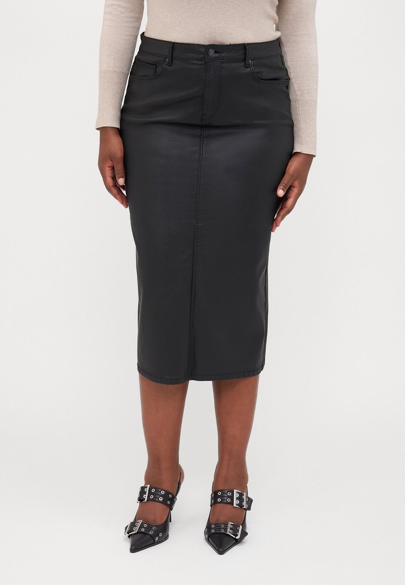 Femme portant une jupe en cuir noire jusqu'aux genoux, un haut beige à manches longues et des chaussures noires pointues avec des boucles argentées.