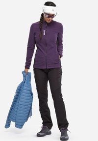 Purple fleece jacket with a zip, black pants, and a light blue insulated jacket held in hand. Gray hiking boots with purple accents.