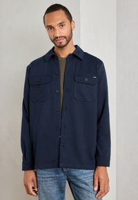 Man wearing navy blue button-up shirt jacket with flap pockets and light blue jeans against a neutral gray background.
