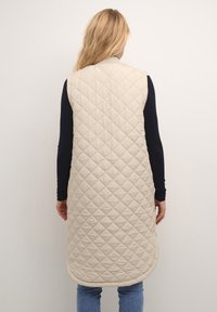 Beżowa pikowana kamizelka o długim kroju. Posiada diamentowy wzór, okrągły dekolt i otwory na ramiona. Nosi się ją na granatowej bluzce z długim rękawem.