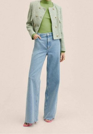 Femme portant un haut côtelé vert clair, une veste en tweed verte raccourcie avec des boutons dorés, un jean bleu taille haute à jambes larges, et des chaussures pointues roses.