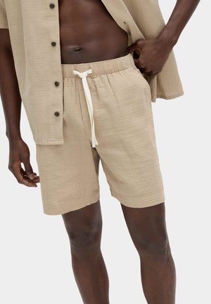 Man draagt beige linnen shorts met een witte trekkoord, gecombineerd met een open beige overhemd met korte mouwen tegen een witte achtergrond.