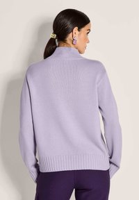 Frau mit dunklen Haaren im Pferdeschwanz, die lila quadratische Ohrringe, einen helllila Rollkragenpullover und dunkelviolette Hosen trägt, von hinten gezeigt.