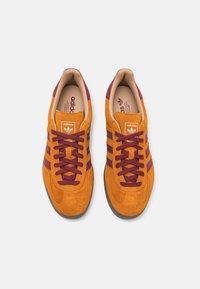 Baskets en daim avec une tige orange, des rayures bordeaux et des lacets assortis ; comporte un logo Adidas classique sur la languette et la semelle intérieure.