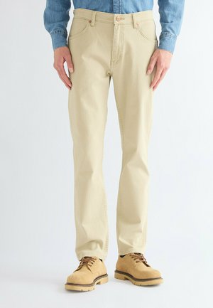 Uomo con pantaloni beige, stivali beige con lacci e camicia blu a maniche lunghe, in piedi con le mani rilassate ai lati.