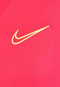 Tejido de camiseta Nike de color rosa con una textura suave, que presenta un logo de swoosh bordado en amarillo en el lado izquierdo.