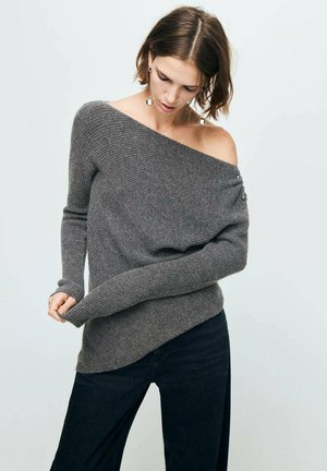 Suéter gris sin mangas con textura acanalada, mangas largas y detalles de botones. Combinado con jeans oscuros de pierna recta.