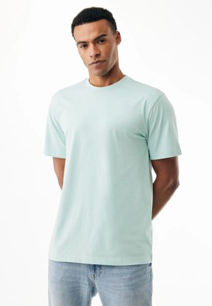 Hombre de pie con las manos detrás de la espalda, vistiendo una camiseta de manga corta azul claro y jeans de lavado claro, frente a un fondo blanco.