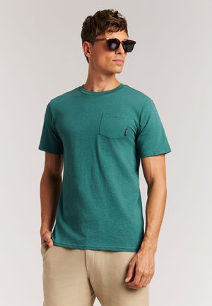 Man draagt een turquoise T-shirt met korte mouwen en borstzak, beige broek, staat met handen in de zakken, draagt donkere zonnebril.