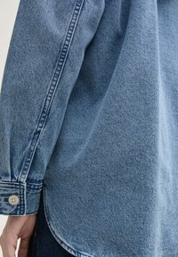Traper košulja u svijetloplavoj boji s mekanom teksturom, koja ima zaobljeni rub, dugmad na manžetama i vidljive šavove duž šavova.