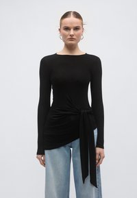 Cămașă neagră cu mâneci lungi, tiv asimetric și detaliu de legare laterală. Material fin, gât rotund, asortată cu blugi largi, de culoare albastru deschis.