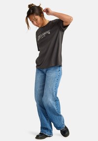 Camiseta gris oscuro con el logo de Billabong, combinada con jeans acampanados de color azul claro y zapatos negros. La tela parece suave con un ajuste relajado.