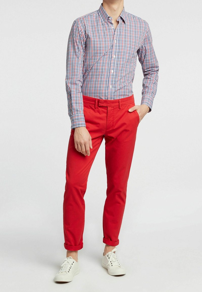 Homme portant des baskets blanches, un pantalon rouge vif retroussé aux chevilles et une chemise à manches longues à carreaux avec des motifs bleus, rouges et blancs.