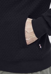Svarte teksturerte genser med ribbestrikkede mansjetter og nederkant, med en sidelomme og subtile stripete detaljer på lomma.
