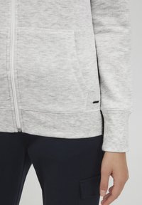Helle graue Zip-Hoodie mit texturierter Oberfläche, Kängurutasche und gerippten Bündchen. Begleitet von dunkelblauen Hosen mit einer Seitentasche.