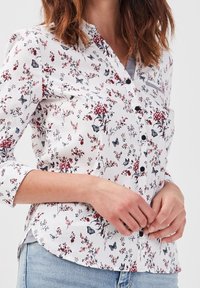 Chemise blanche à boutons, ornée d'un imprimé floral et de papillons en rouge et gris, avec un ourlet arrondi et des manches retroussables.