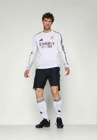 Witte lange mouwen voetbaltrui met zwarte accenten en het logo "Emirates Fly Better", gecombineerd met zwarte shorts en witte sokken met strepen.