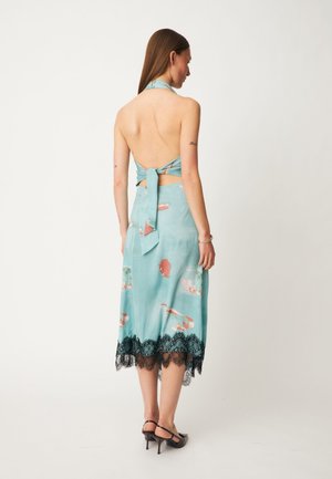 Femme portant une robe dos nu halter bleu clair à imprimés floraux et ourlet en dentelle noire, debout et dos tourné sur un fond uni.