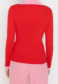 Roter, gerippter Pullover mit langen Ärmeln und einem kontrastierenden rosa Kragen. Die Rückseite ist tailliert gestaltet und zeigt eine glatte Textur.