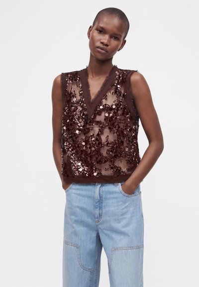 Femme portant un top sans manches marron foncé transparent avec des sequins et un jean large bleu clair, debout les mains dans les poches.