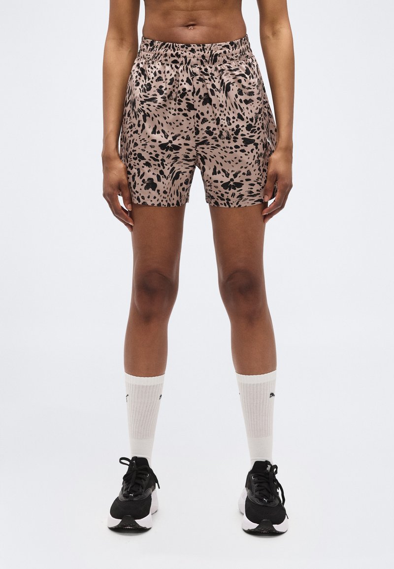 Άτομο που φοράει σορτς μπεζ με μαύρες βούλες, λευκές κάλτσες μέχρι τον αστράγαλο και μαύρα αθλητικά παπούτσια, στέκεται μπροστά σε ένα απλό φωτεινό φόντο.